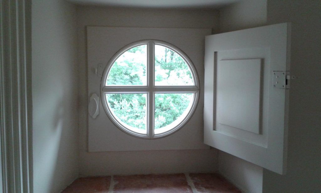Petite pièce avec une fenêtre ronde et une porte blanche ouverte, laissant apparaître la verdure à l’extérieur.