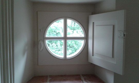Petite pièce avec une fenêtre ronde et une porte blanche ouverte, laissant apparaître la verdure à l’extérieur.