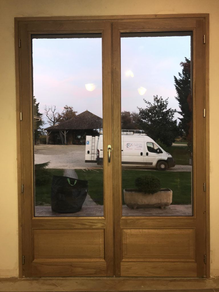 Portes doubles en bois avec des vitres donnant sur une rue, une camionnette et un ciel nuageux en soirée.