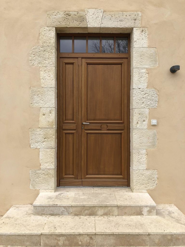 Porte d’entrée en bois avec encadrement en pierre et petite marche, encastrée dans un mur en stuc beige.