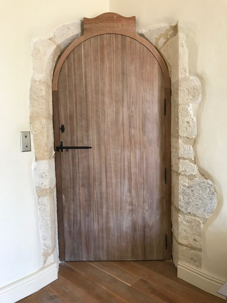 Porte cintrée en bois encastrée dans une embrasure en pierre, avec une poignée noire, vue de l’intérieur sur un parquet en bois à chevrons