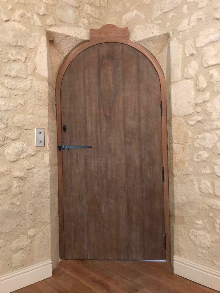 Porte cintrée en bois encastrée dans un mur de pierre, avec une poignée argentée et un interrupteur à côté.