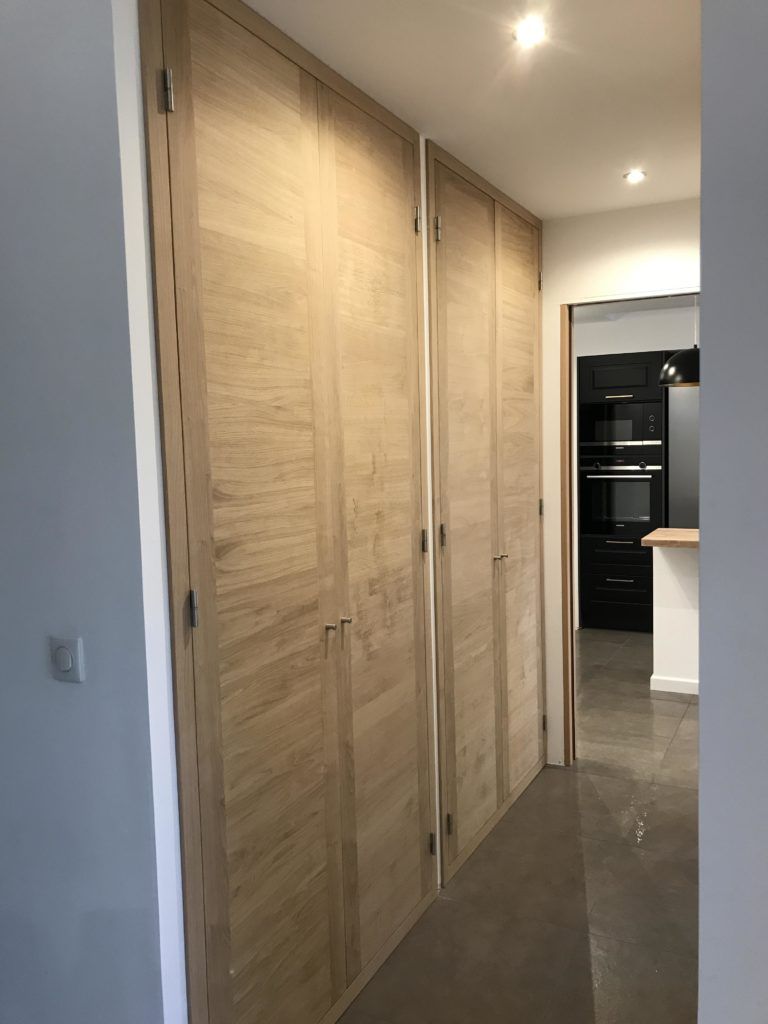 Couloir avec de hautes portes de placard en bois clair et des spots encastrés au plafond, menant à une cuisine.