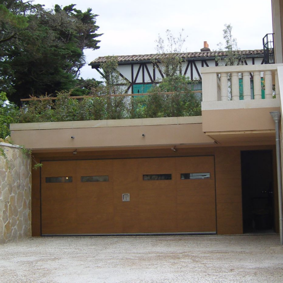 Extérieur d’une maison moderne avec une porte de garage en bois, un mur en pierre et un balcon donnant sur une allée