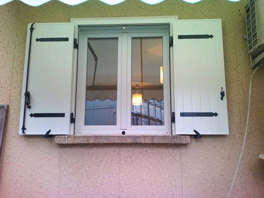Fenêtre aux volets blancs sur un mur extérieur beige, avec une lampe allumée visible à l’intérieur à travers la vitre