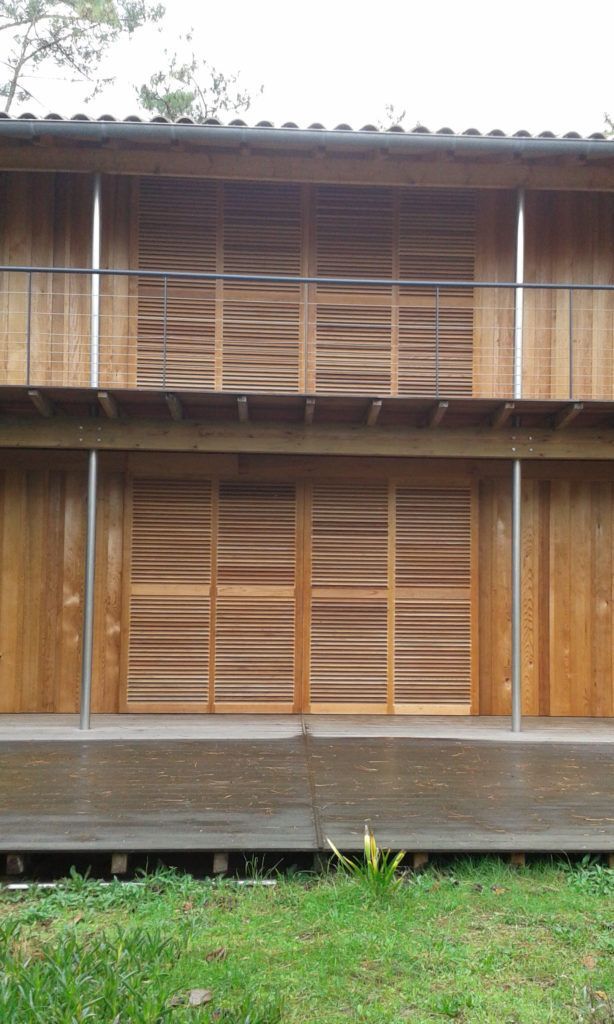 Maison en bois à deux étages, avec des fenêtres à persiennes et un petit jardin verdoyant devant.