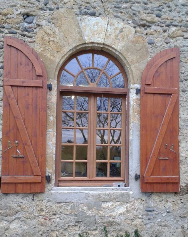 Mur en pierre avec une fenêtre cintrée et des volets en bois brun ouverts
