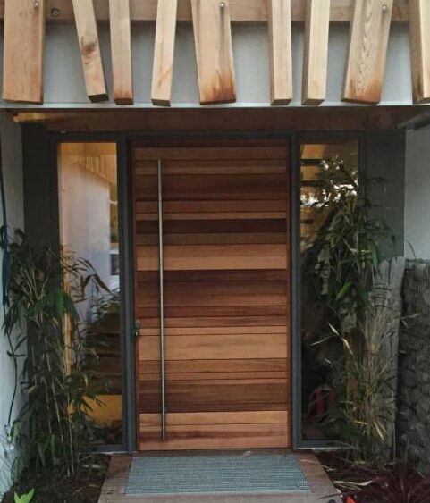 Porte d’entrée moderne en bois à panneaux verticaux, encadrée de plantes et d’une entrée en pierre