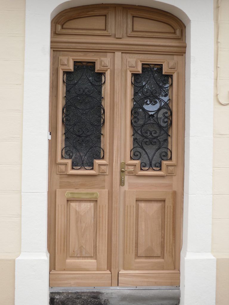 Deux portes d’entrée en bois ornées de panneaux en fer forgé noir et en verre, surmontées d’une imposte cintrée.