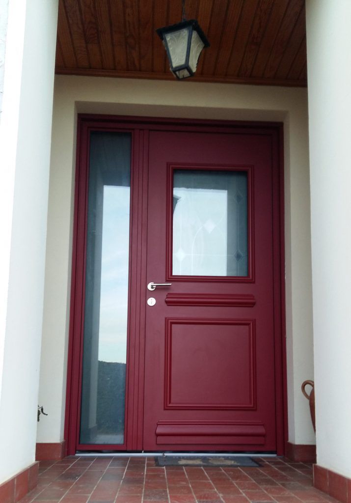 Porte d’entrée rouge à panneaux vitrés sous une applique extérieure, encadrée de murs blancs et de marches en briques.