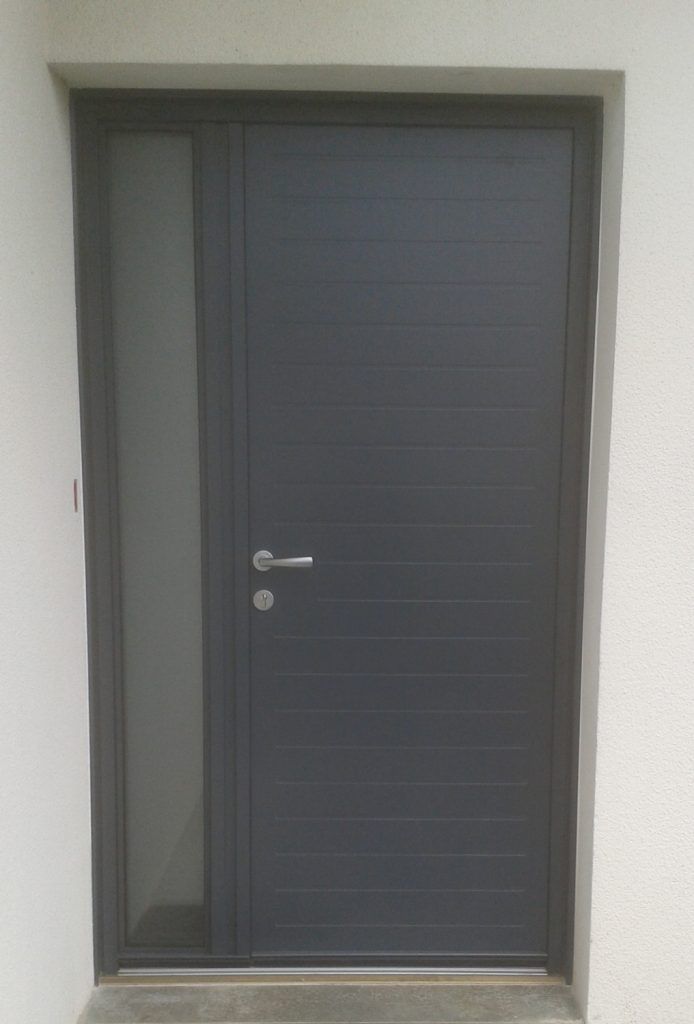 Porte d’entrée grise avec une poignée argentée, encastrée dans un mur blanc, et une fenêtre latérale étroite