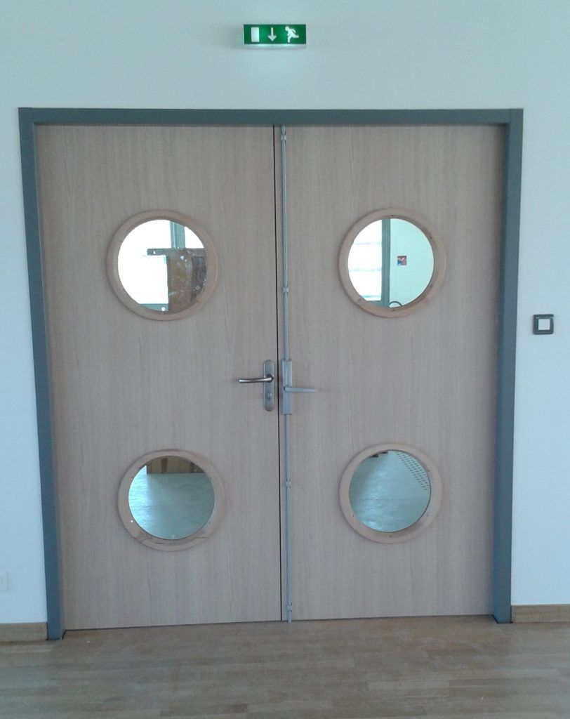 Portes doubles en bois avec des hublots ronds dans un couloir bleu clair, avec un panneau de sortie au-dessus.