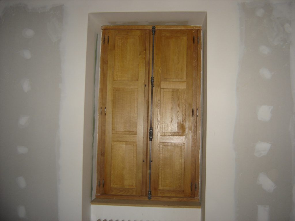 Volets en bois à deux vantaux encastrés dans une niche d’un mur blanc, avec des zones d’enduit non finies autour