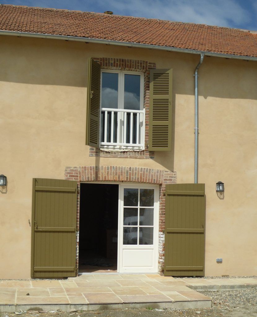 Façade beige avec des volets ouverts, une fenêtre donnant sur un balcon et une porte blanche ouverte.