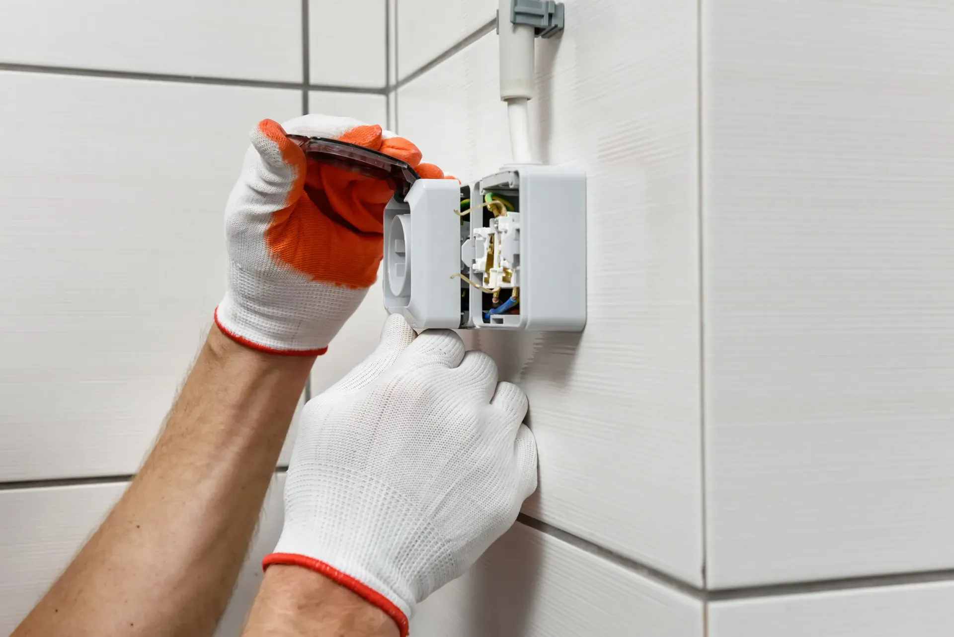 Persona con guantes, trabajando en una caja eléctrica blanca sobre una pared de azulejos.