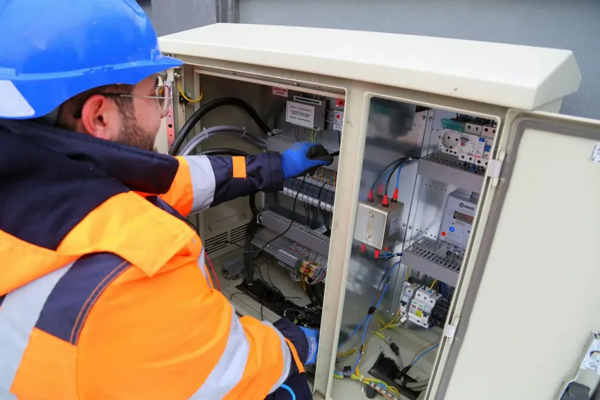 Persona con ropa de trabajo naranja y azul y casco trabajando en un panel eléctrico.