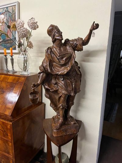 Scultura in legno raffigurante un uomo con turbante che regge un vaso e tiene le braccia tese su un piedistallo.