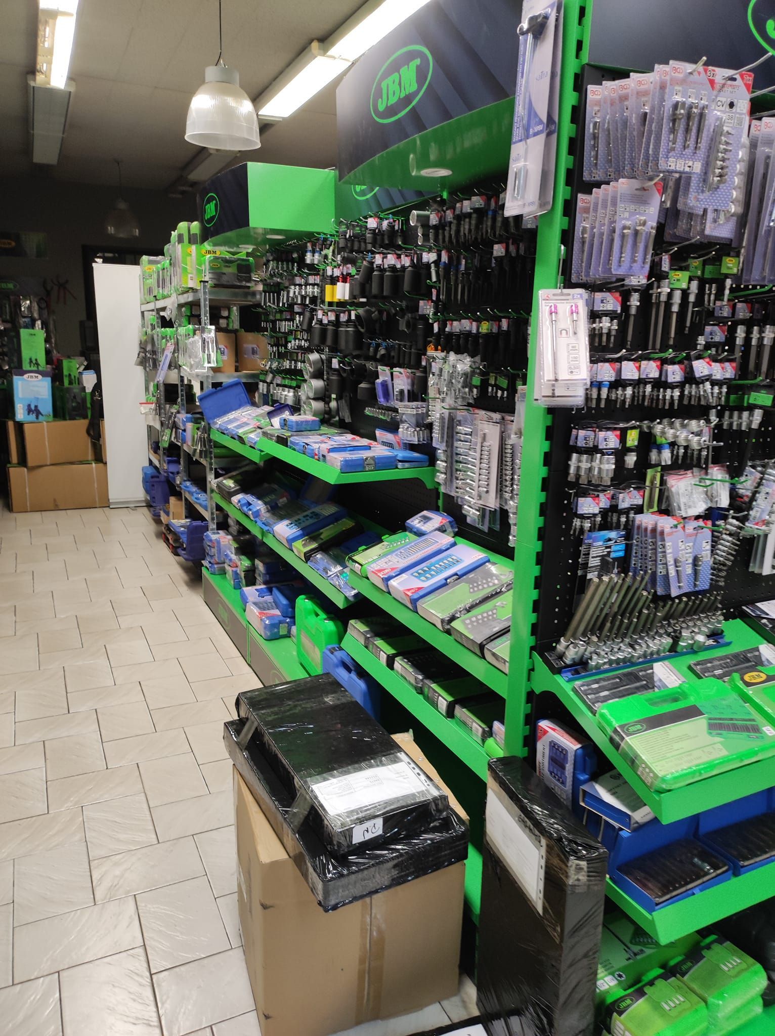 Une allée de vente au détail avec des étagères vertes remplies de divers outils et articles de quincaillerie, menant vers le fond du magasin.