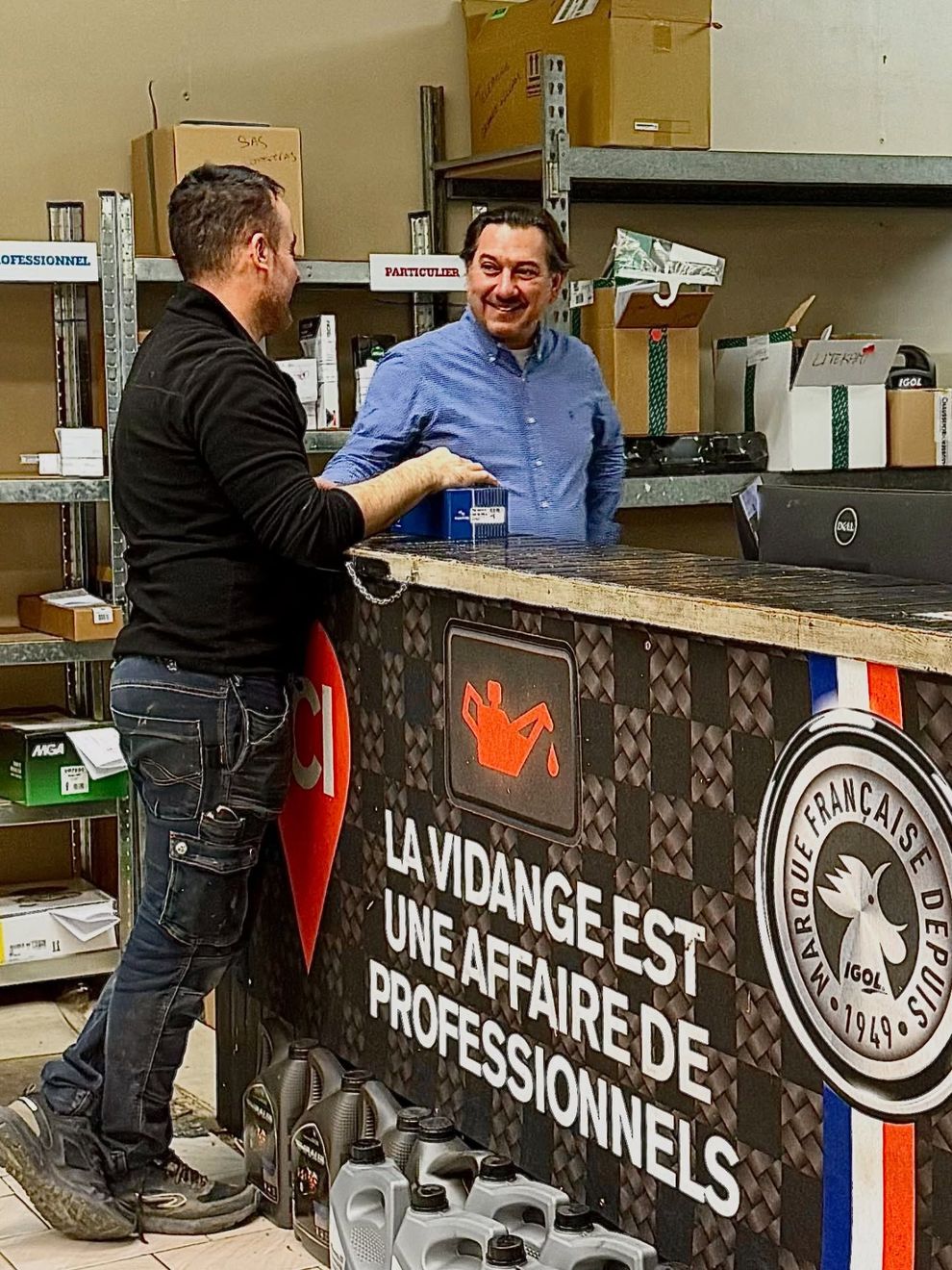Un employé du service d'assistance d'un entrepôt parle avec un client ; le comptoir indique : La vidange est une affaire de pro.