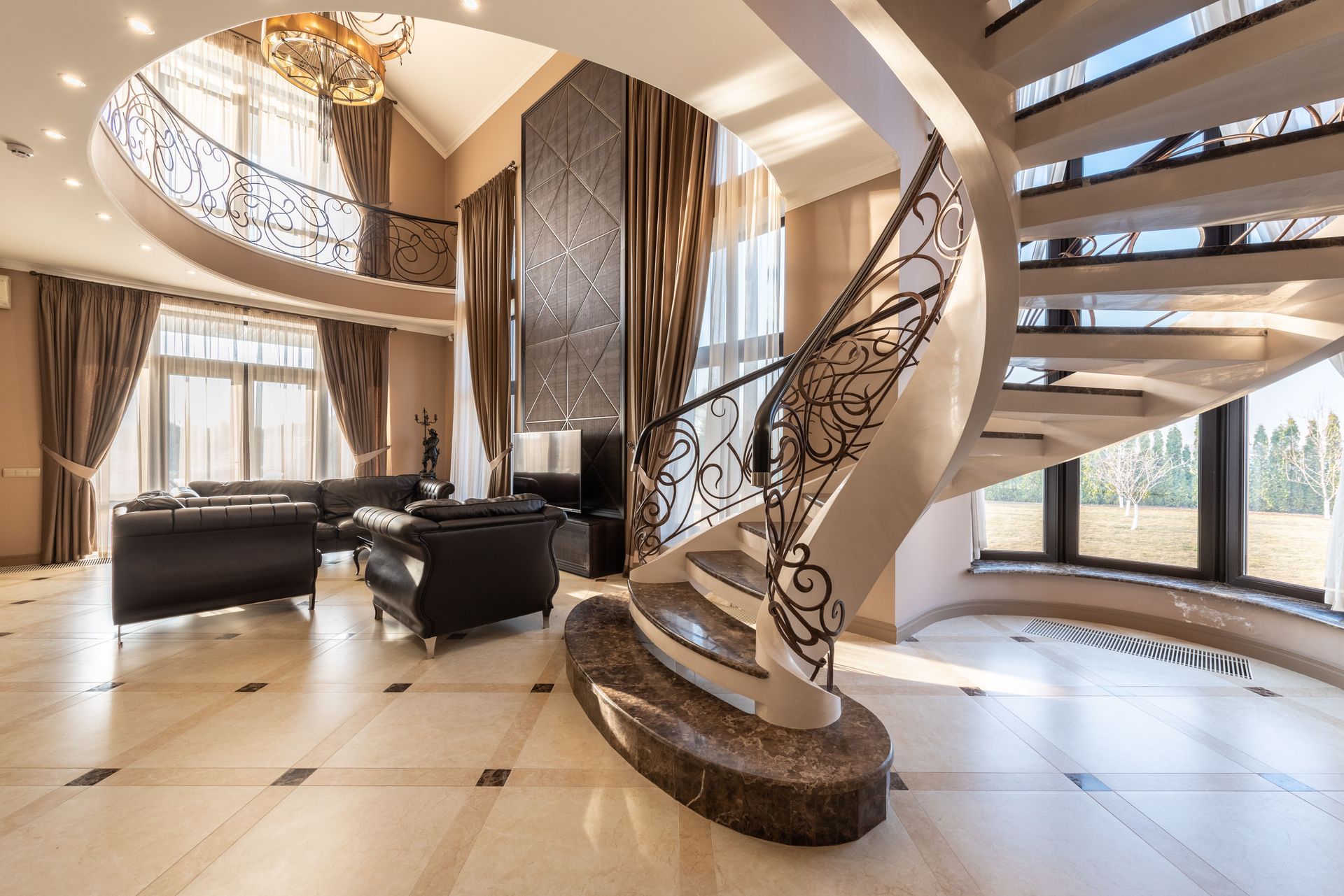Luxurious open foyer with curved staircase, balcony, large windows, and dark leather seating area