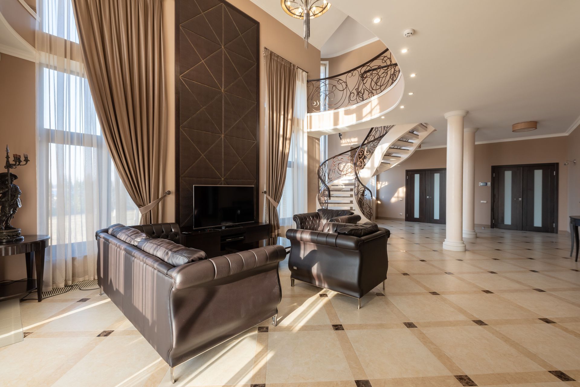 Luxury living room with leather sofas, grand spiral staircase, and tall windows with drapes