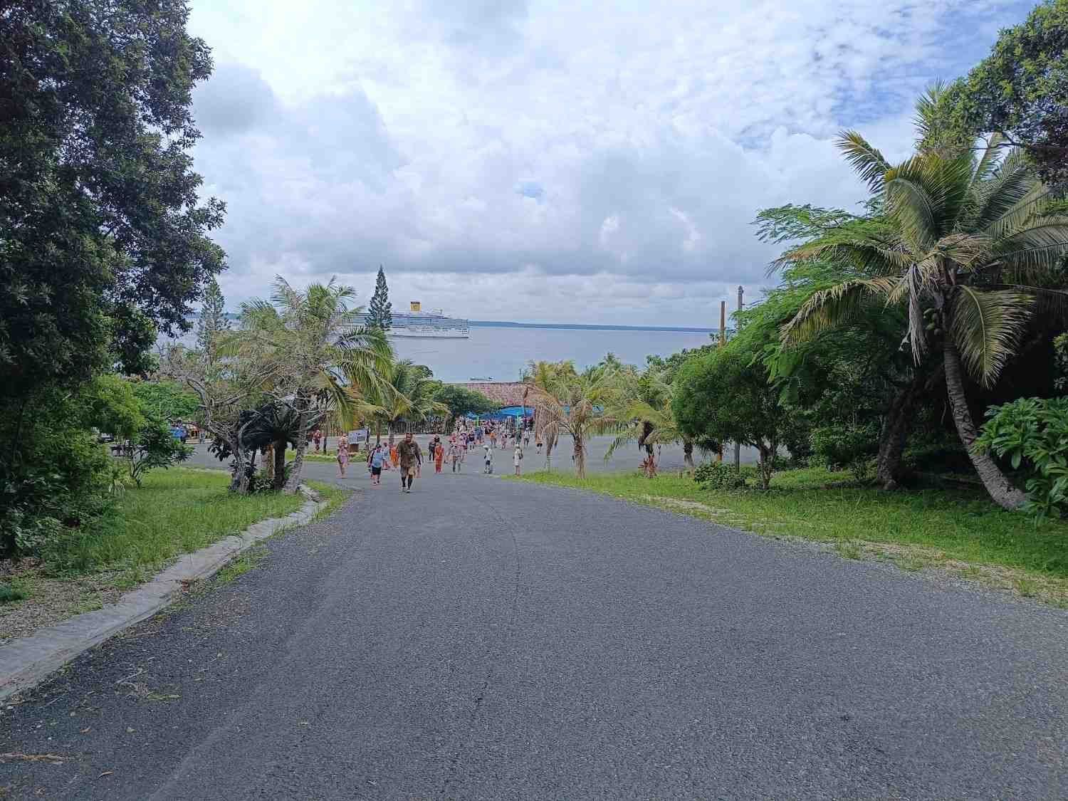Gente caminando por un camino pavimentado hacia la orilla del mar, bordeado de árboles verdes bajo un cielo nublado.