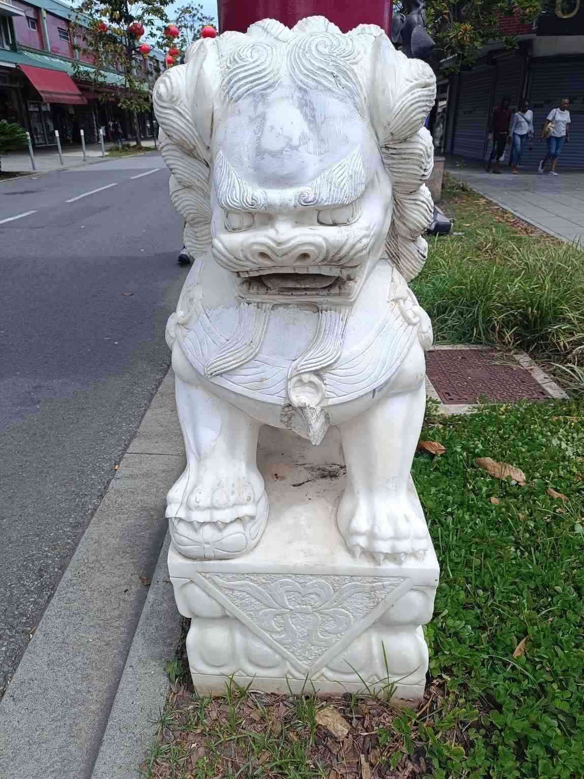 Estatua de un león guardián de piedra blanca en una acera junto al césped y una calle.