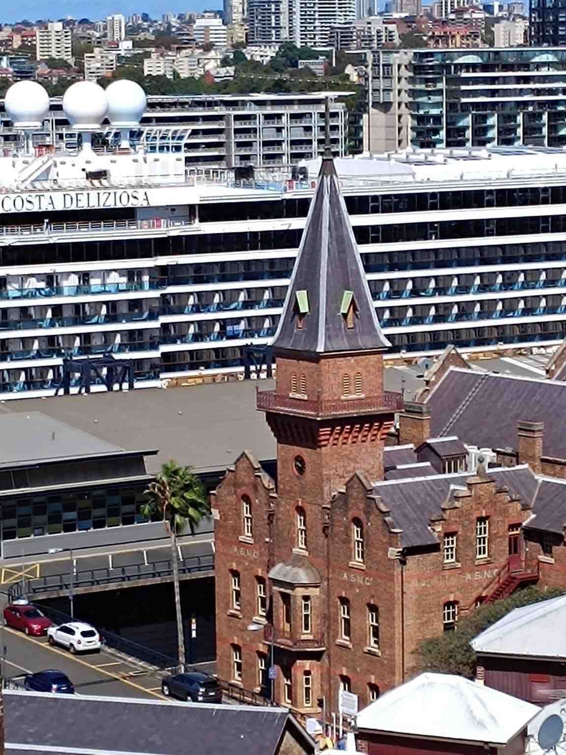 Torre de iglesia de ladrillo rojo frente a un gran crucero atracado en el paseo marítimo.