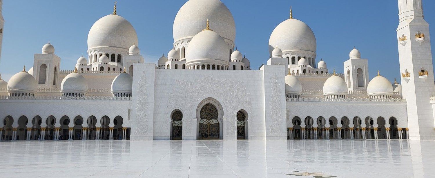 Gran mezquita blanca con cúpulas y minaretes que se reflejan en un patio resplandeciente.