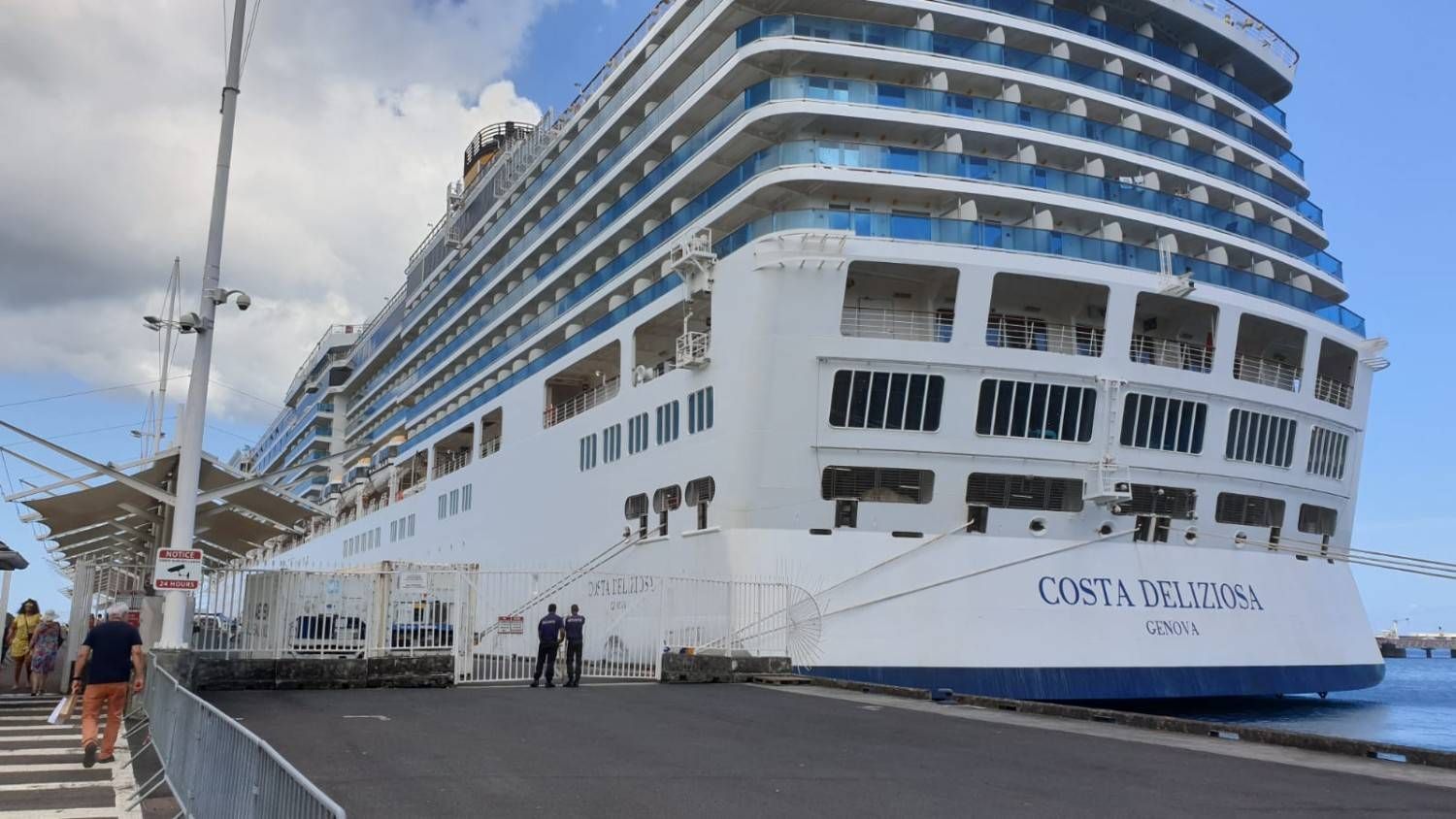 Un gran crucero blanco atracó en un puerto, con gente caminando por el muelle bajo un cielo nublado.
