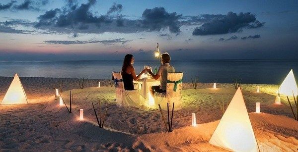 Gente cenando a la luz de las velas en la playa al atardecer, con faroles brillantes y las olas del océano de fondo.