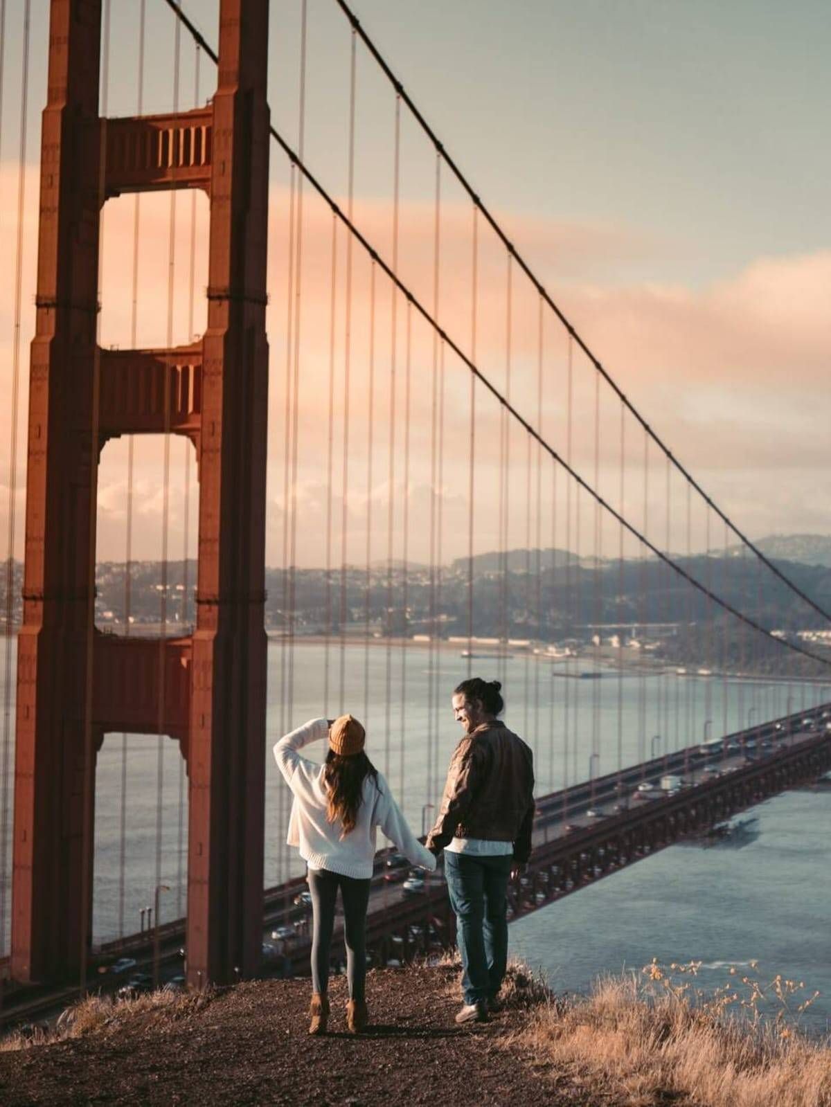 Dos personas se encuentran de pie cerca del puente Golden Gate al atardecer, contemplando la bahía.