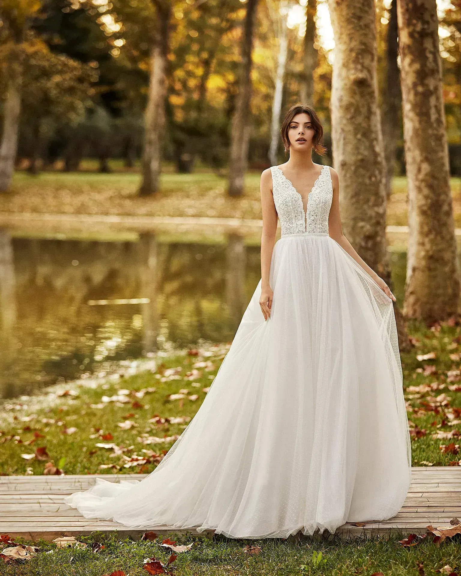 Una mujer con un vestido de novia blanco está parada en un muelle de madera frente a un lago.