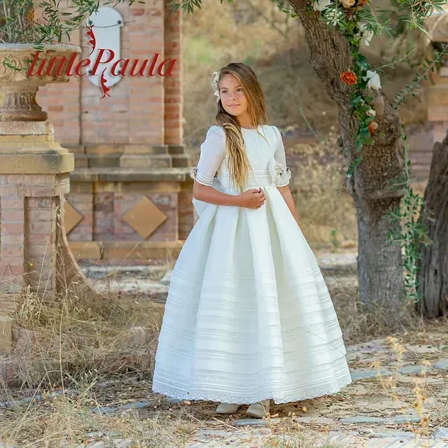 Una niña con un vestido blanco está parada frente a un árbol.