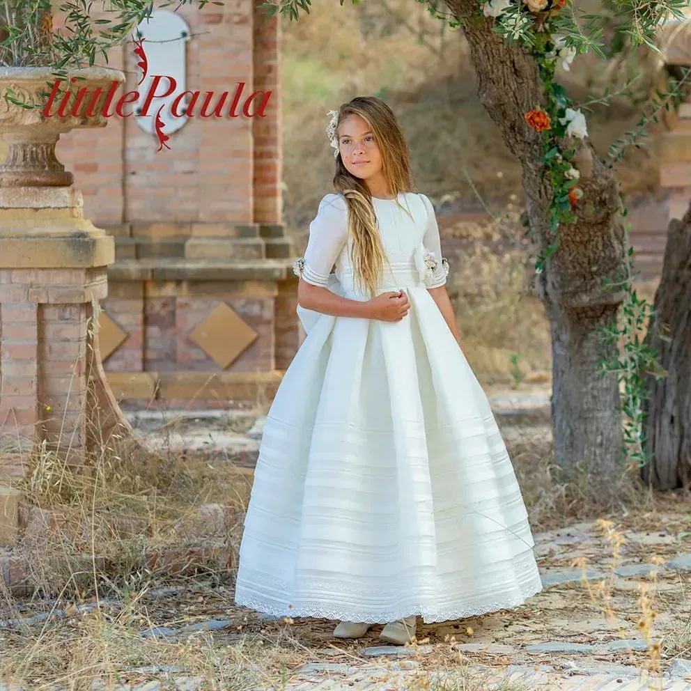 Una niña con un vestido blanco está parada frente a un árbol.