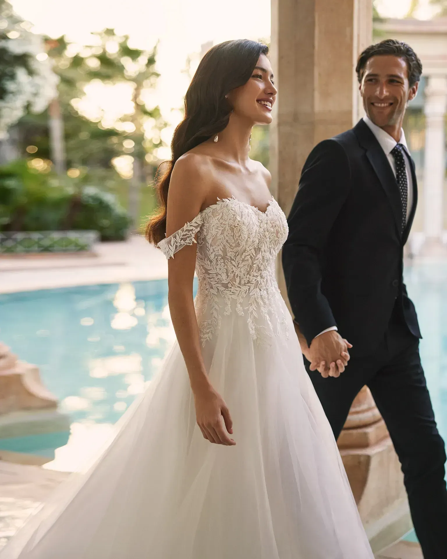 Una novia y un novio se toman de la mano mientras caminan al lado de una piscina.