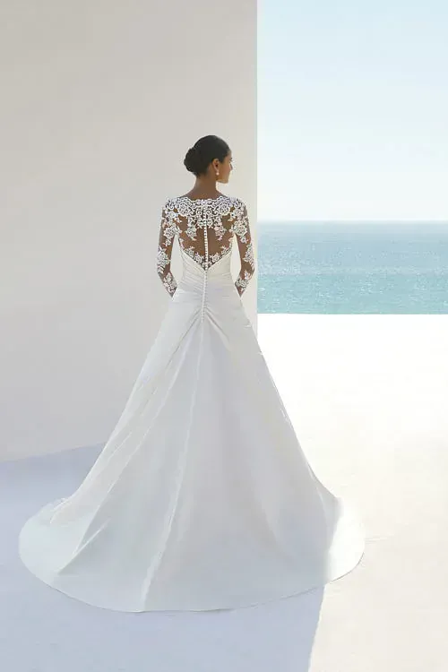 Una mujer con un vestido de novia blanco está parada frente a una ventana con vista al océano.