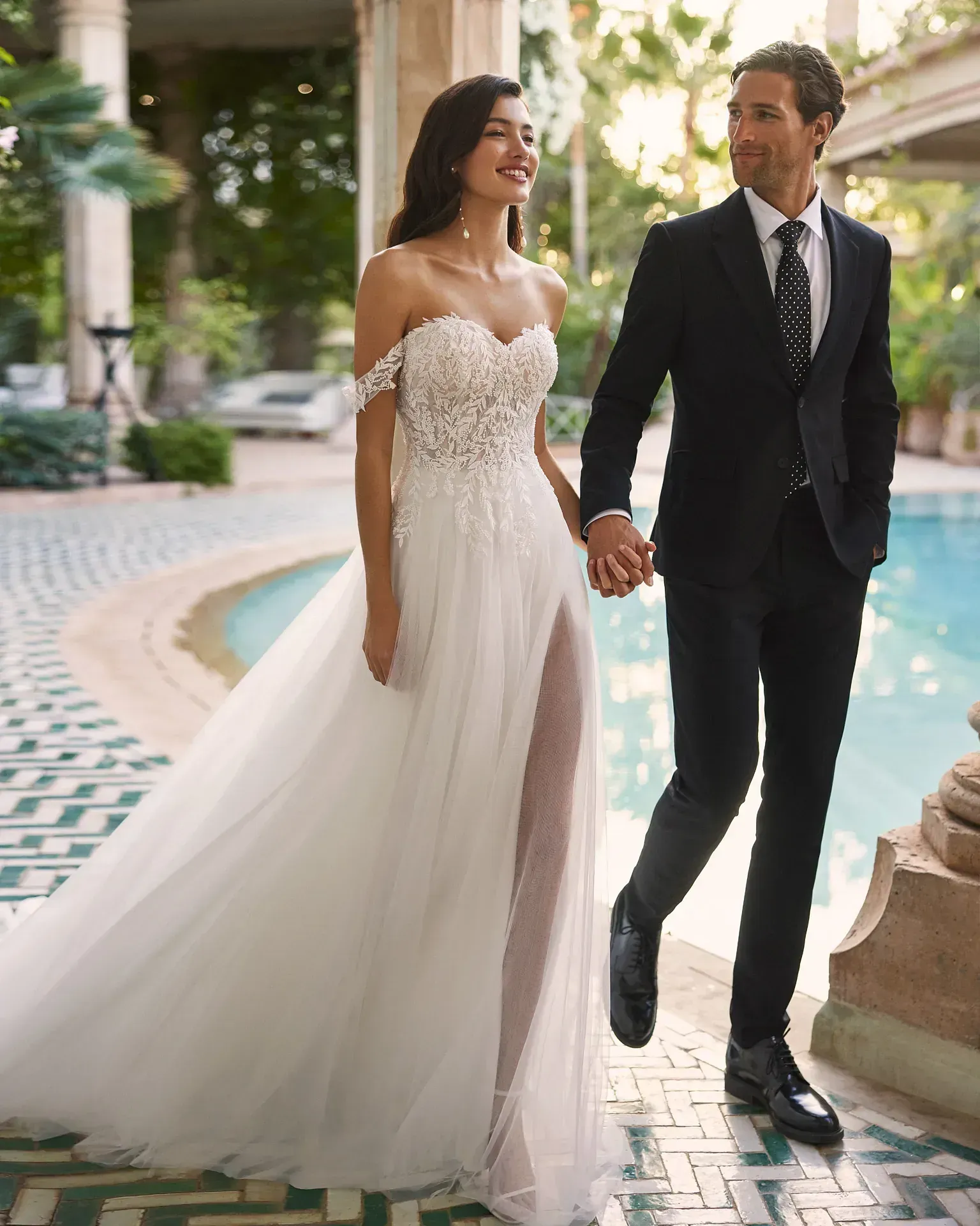 Una novia y un novio se toman de la mano mientras caminan junto a una piscina.