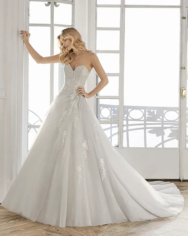 Una mujer lleva un vestido de novia blanco y está parada frente a una ventana.