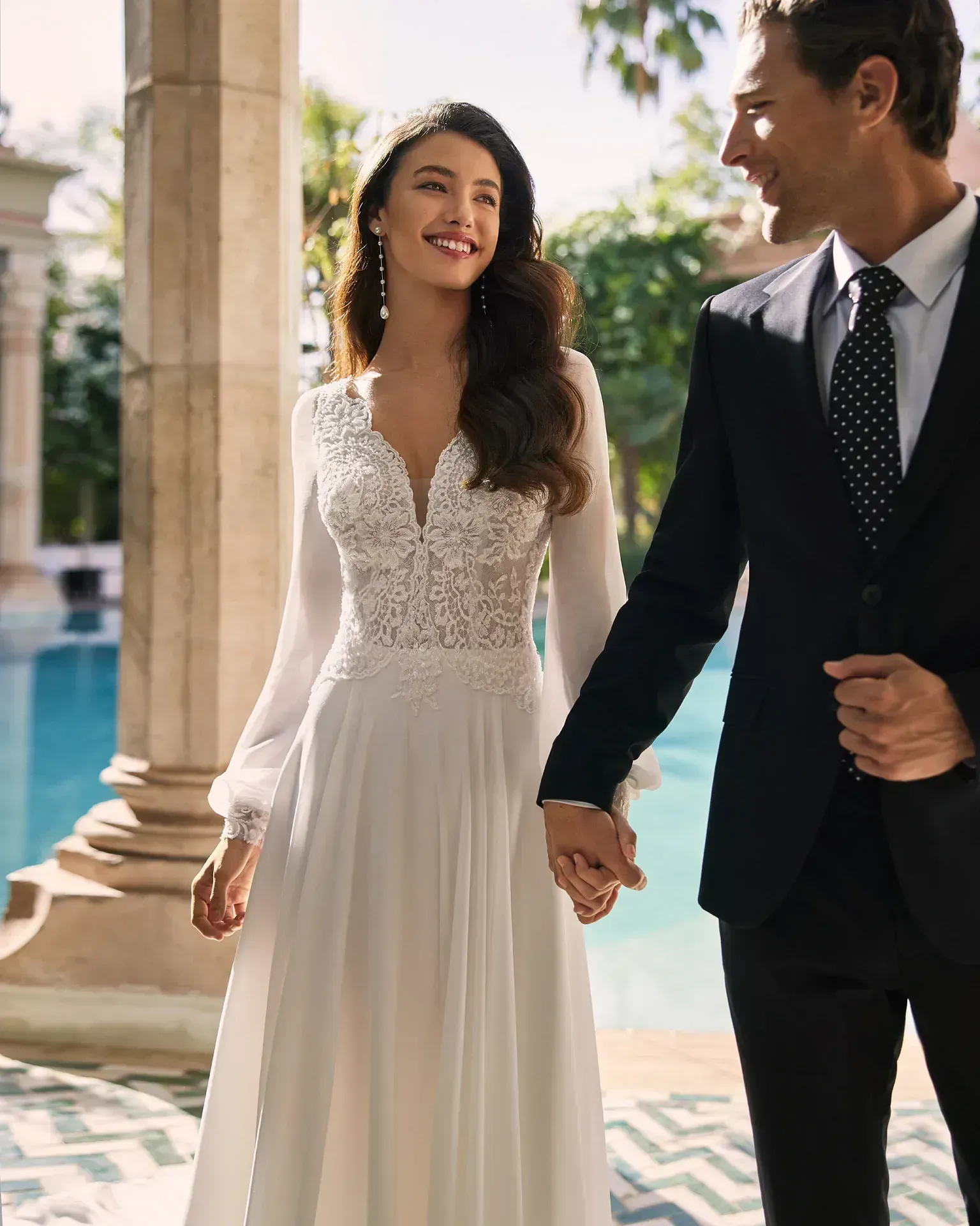 Una novia y un novio se toman de la mano mientras están de pie junto a una piscina.