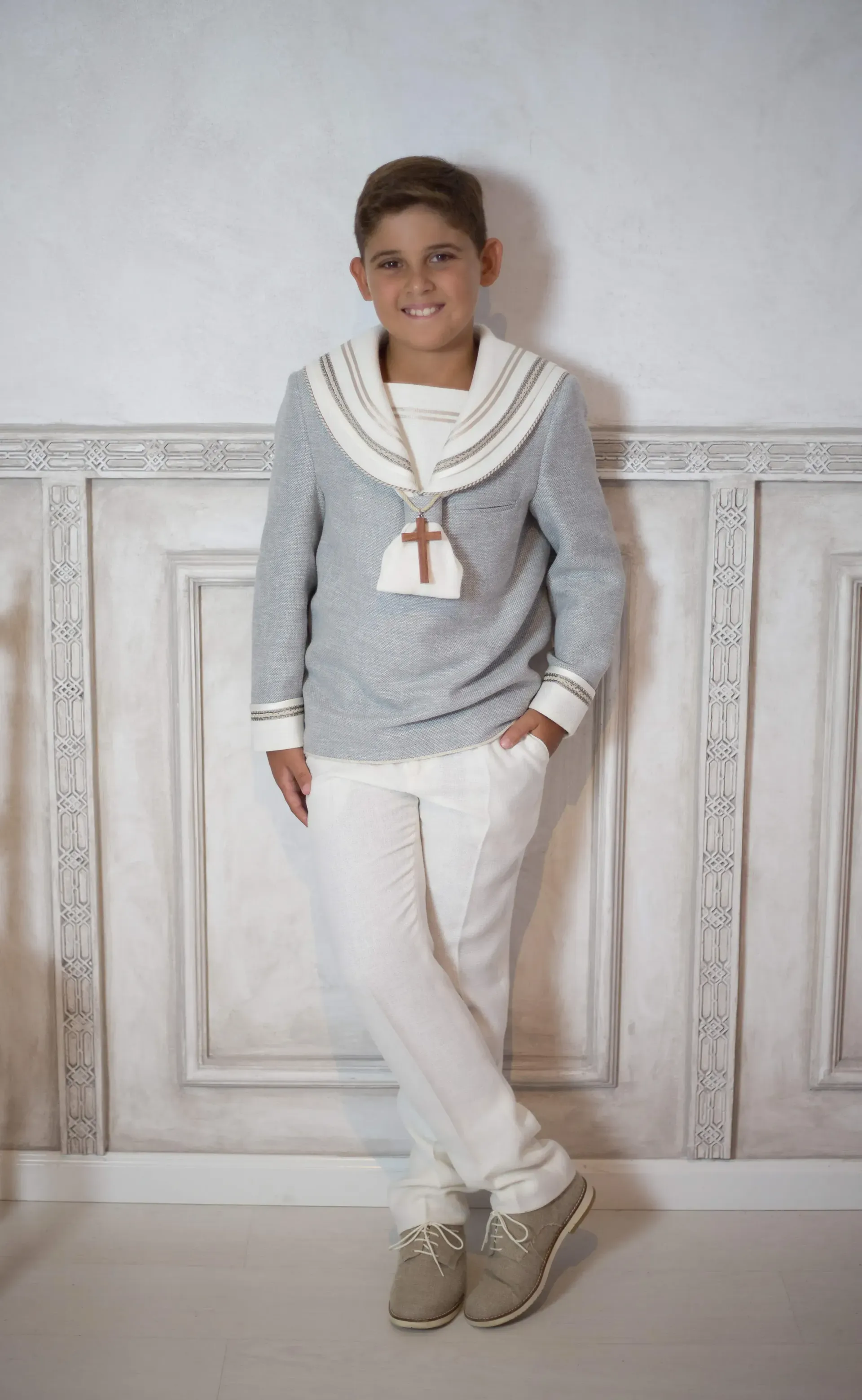 Un niño pequeño con un suéter y pantalones blancos está parado frente a una pared blanca.