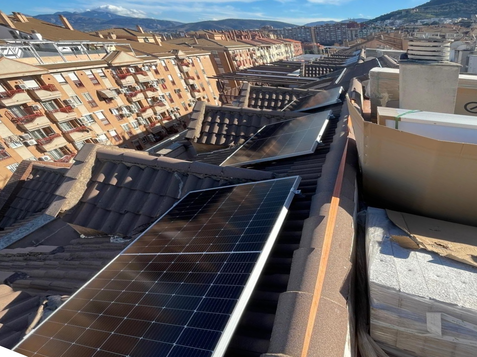 Vista desde la azotea de paneles solares dañados y tejado de tejas con edificios al fondo. Montañas y cielo azul.