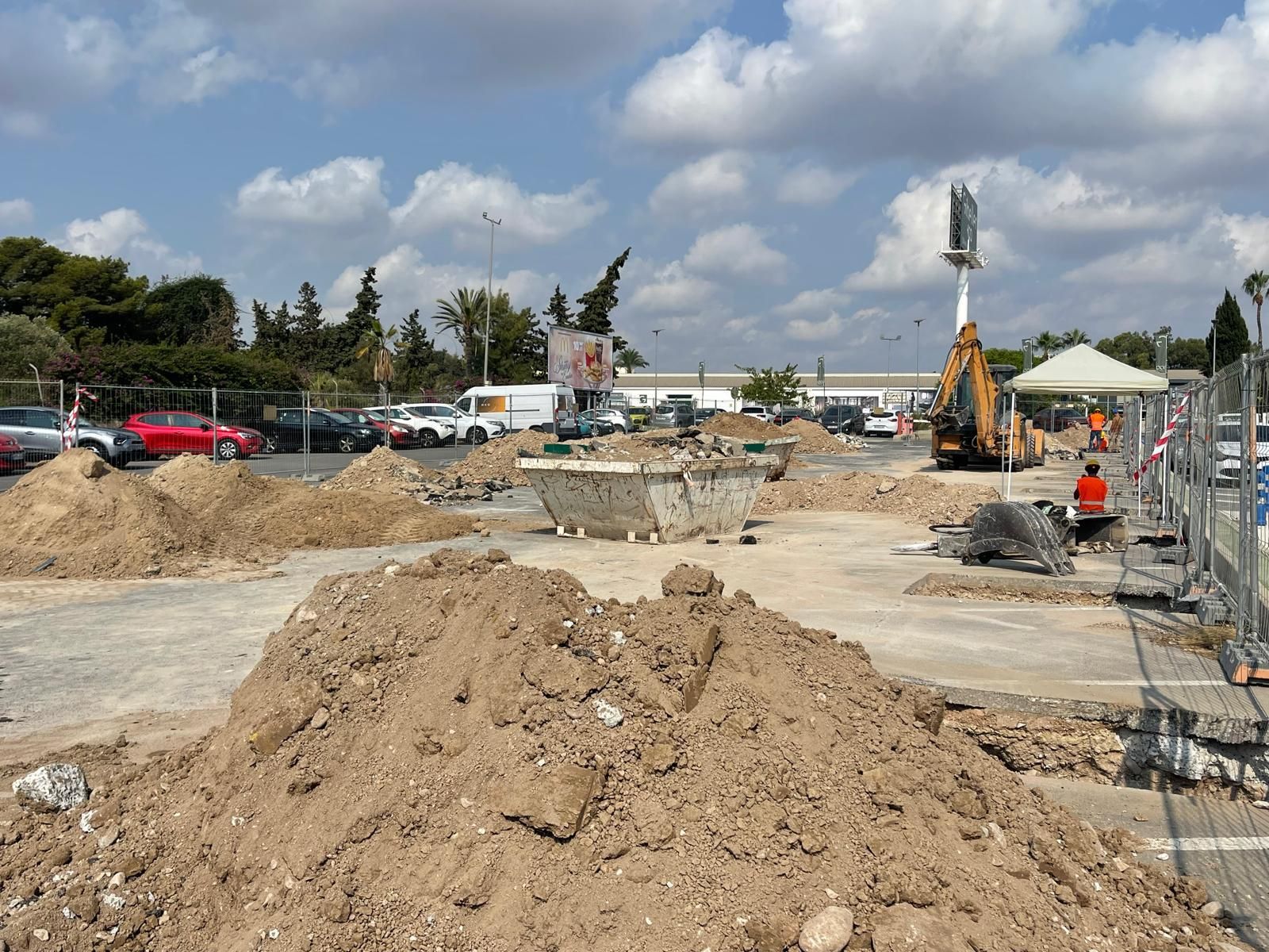 Sitio de construcción: montones de tierra, maquinaria, área de concreto parcialmente construida, autos estacionados, cielo azul.
