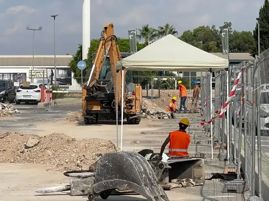 Sitio de construcción: Excavadora, trabajadores con chalecos de seguridad, carpa temporal, escombros de hormigón, cercas.