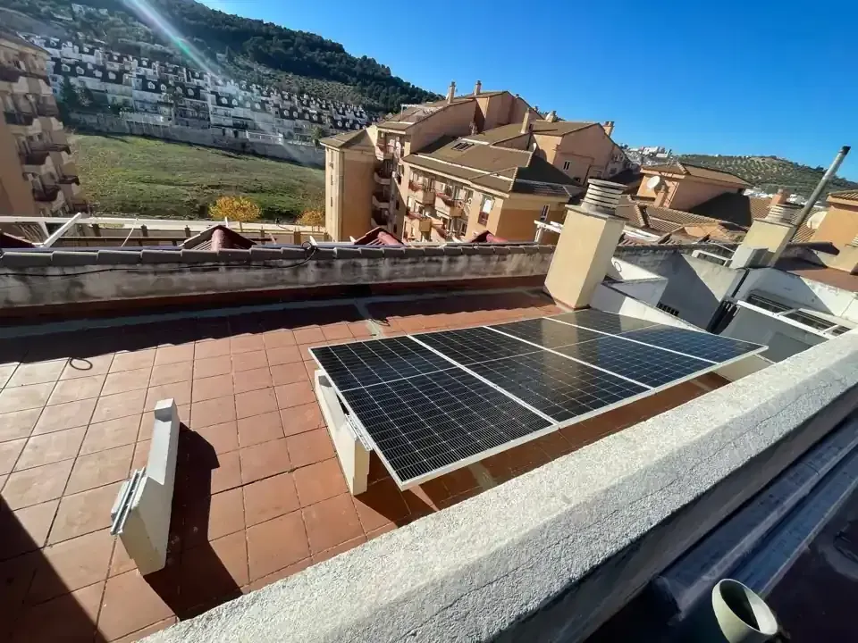 Paneles solares en un tejado, día soleado. Edificios y una ladera visibles al fondo.