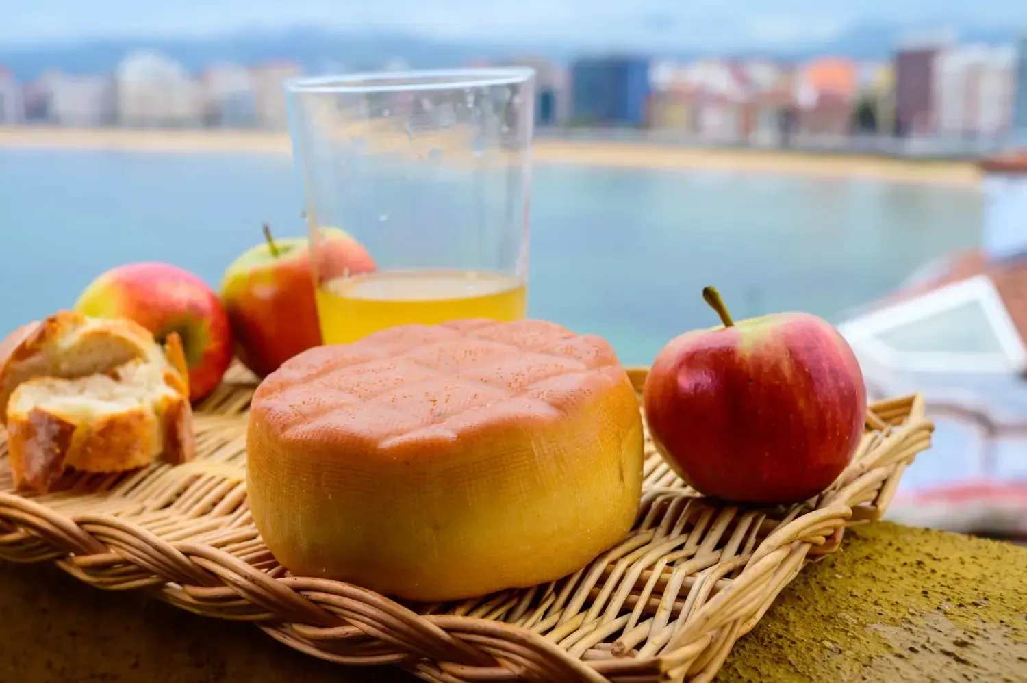 Manzanas, pan, queso y jugo en una bandeja de mimbre con una playa y un paisaje urbano de fondo.