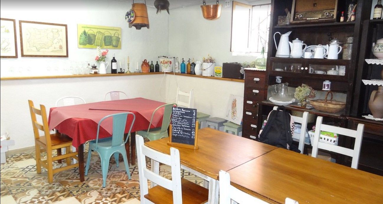 Interior de una cafetería con mesas, sillas y estanterías repletas de artículos. Una mesa tiene un mantel rojo.