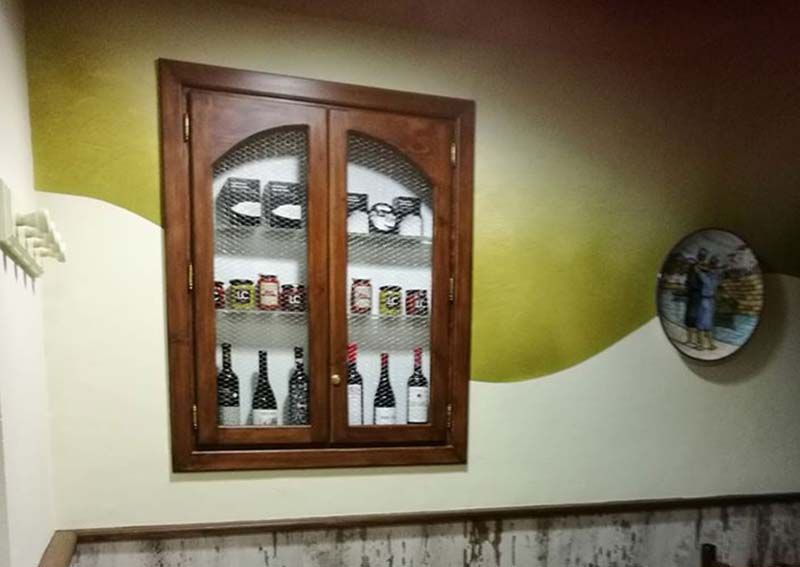 Armario de madera con tapa arqueada, lleno de botellas y frascos, montado en una pared pintada.