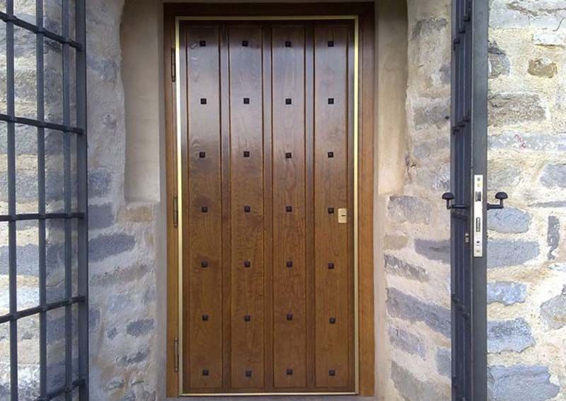 Puerta de madera con paneles verticales e incrustaciones cuadradas, enmarcada en oro contra una pared de piedra.