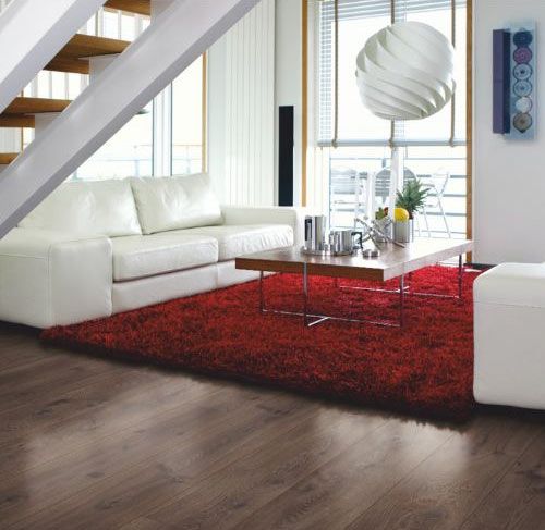 Sala de estar con sofá blanco, alfombra roja, piso de madera y lámpara moderna.